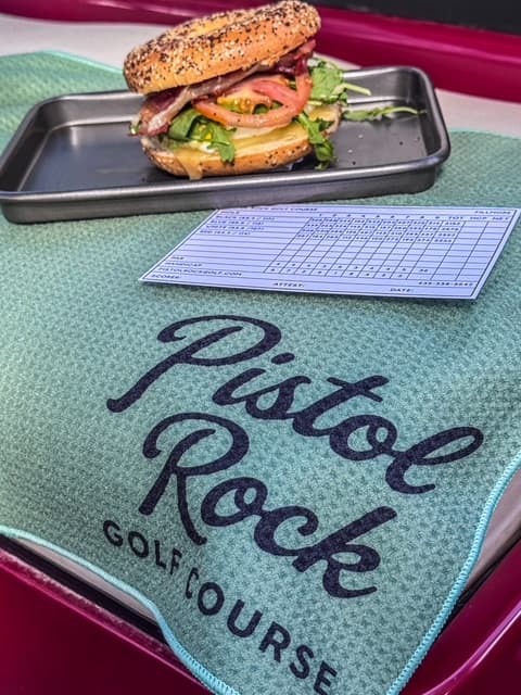 An egg and bacon breakfast sandwich on an everything bagel, next to a Pistol Rock scorecard and golf towel