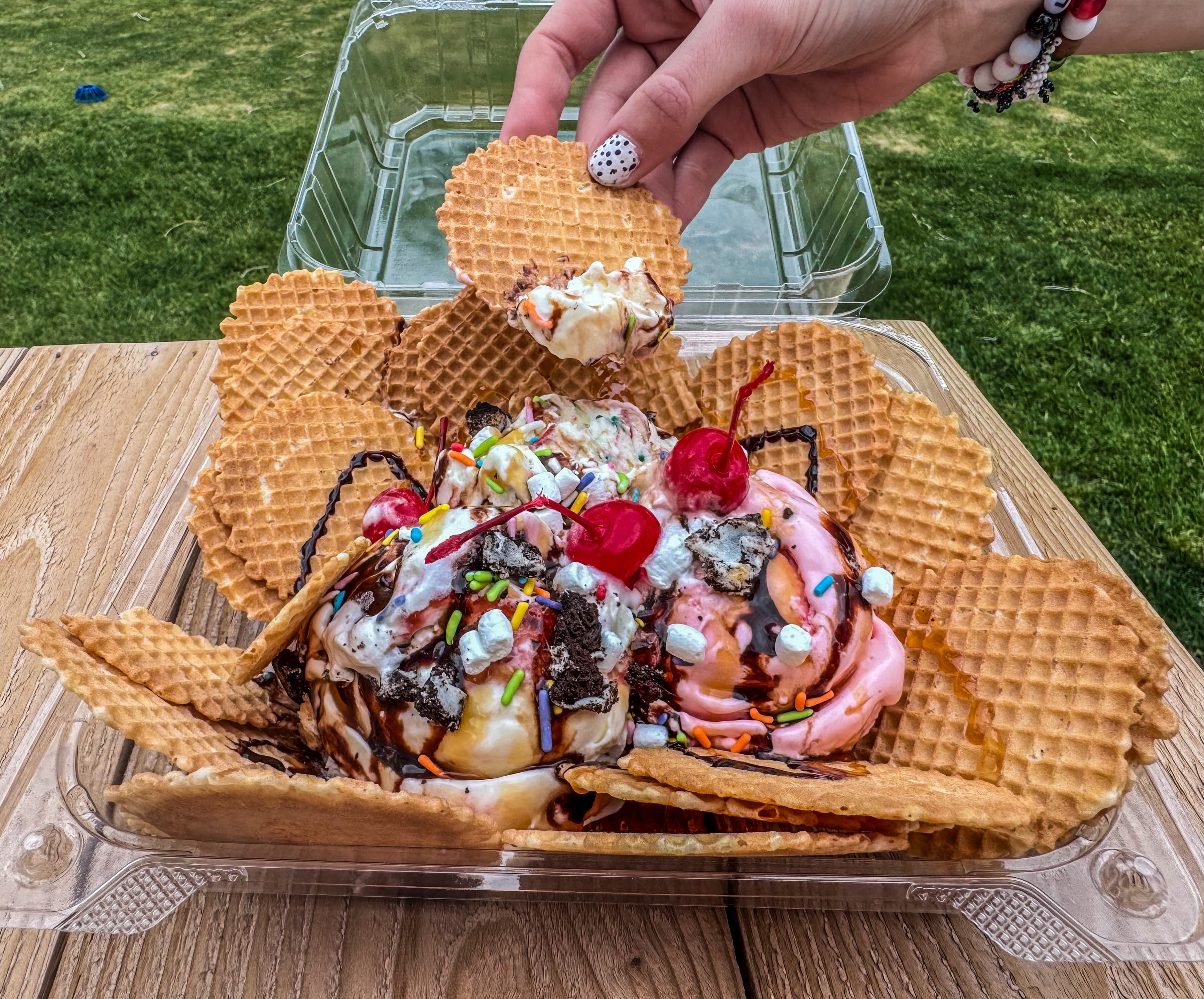 Hand placing a waffle chip onto a sundae of pink and white ice cream with sprinkles and chocolate drizzle on a picnic table